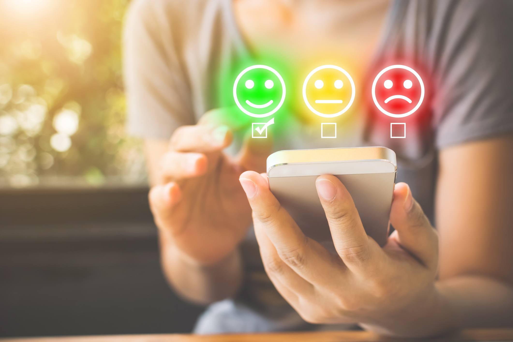 Someone using their phone. Three icons displayed above the phone, a green smiley face, a yellow neutral face and a red sad face.